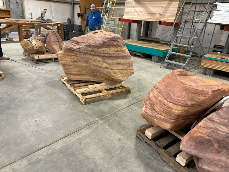 Large, windswept boulders on wooden pallets in an indoor workshop setting, with a person in the background and various tools and equipment visible.