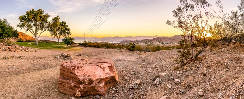 A desert landscape at sunset with a large rock in the foreground, sparse vegetation, and trees in the distance.