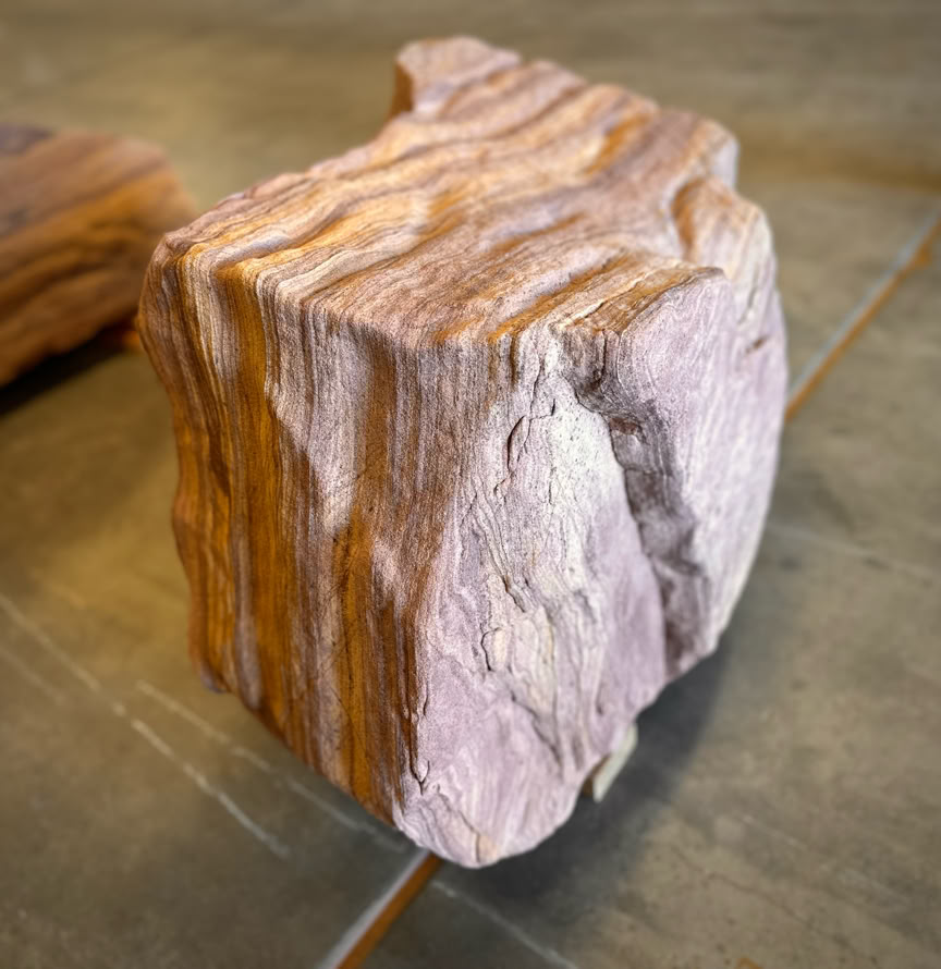 A rough, rectangular piece of rock with layered, striped patterns in shades of brown and beige, placed on a flat surface.