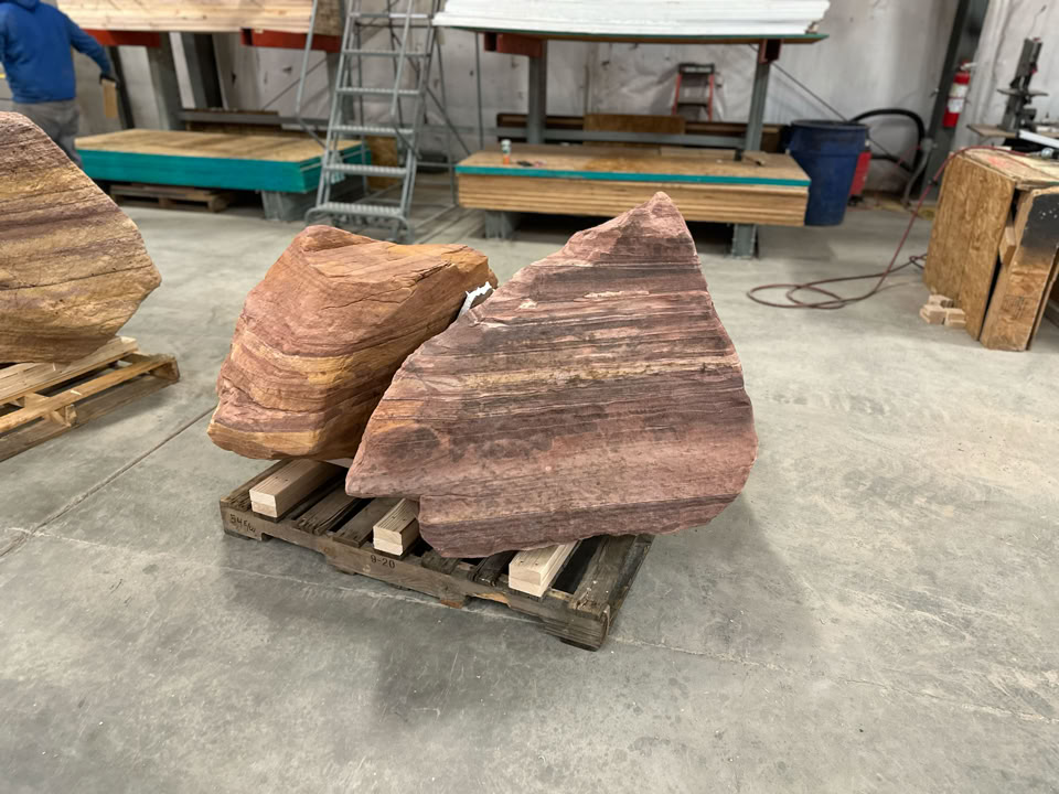 Large, windswept boulders on wooden pallets in an indoor workshop setting, with a person in the background and various tools and equipment visible.