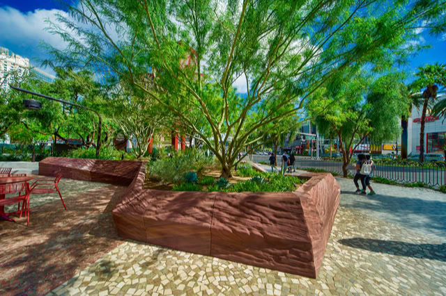 A landscaped area with a large tree and plants in a raised, angular stone planter, surrounded by a paved walkway and people walking in the background. @MGM THE PARK LAS VEGAS NV