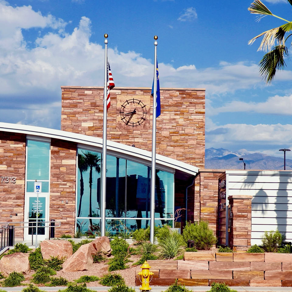 Modern building with stone facade, large glass windows, two flagpoles, and palm trees in front.