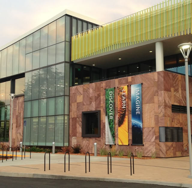 A modern building with large glass windows and a Stone facade, featuring banners and a sign that reads "Library" near the entrance. at Mitchell Park Library