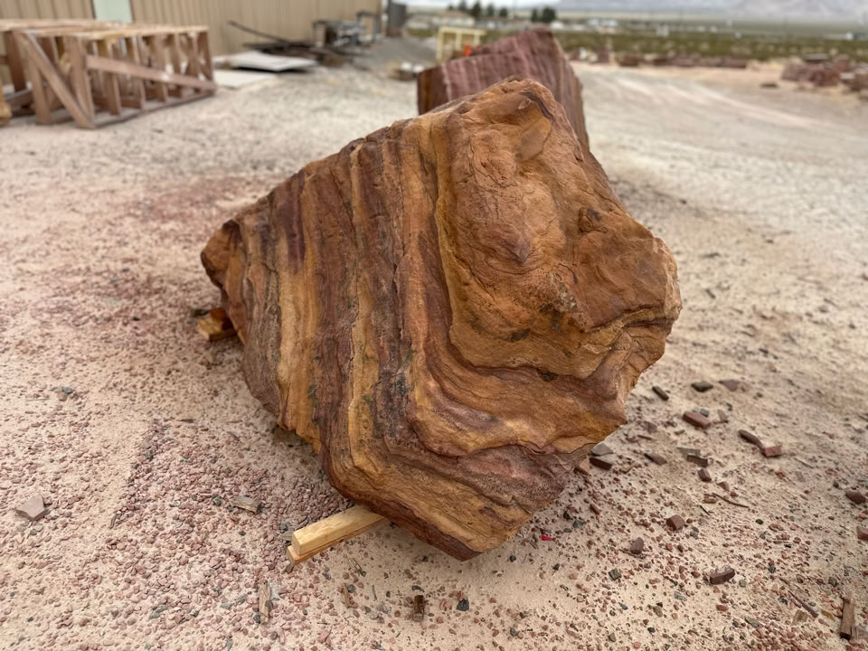 A large, textured rock with brown and orange striations resting on a sandy surface outdoors.
