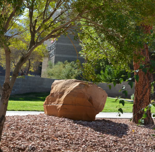 A large rock surrounded by trees and shrubs on a landscaped area with a grassy lawn and a building in the background. at UNLV