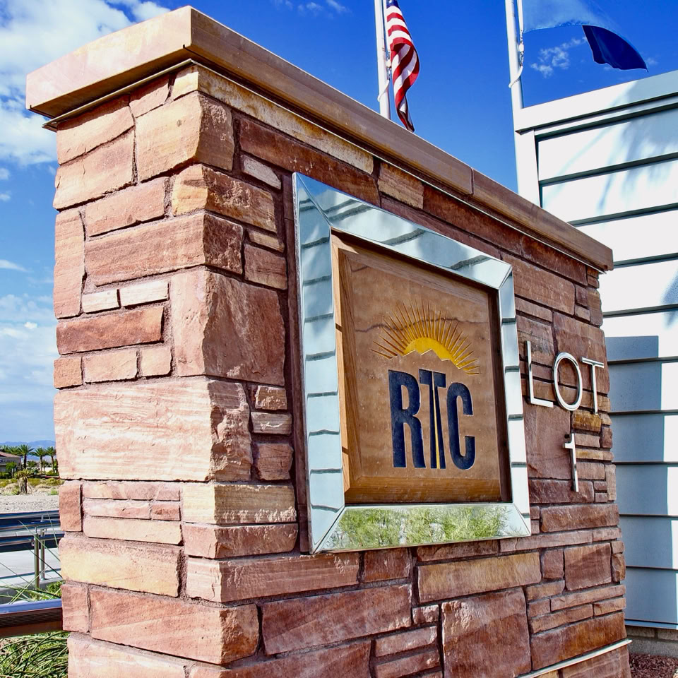 A stone sign with the letters "RTC" and an arrow pointing right, with two flags in the background against a blue sky.