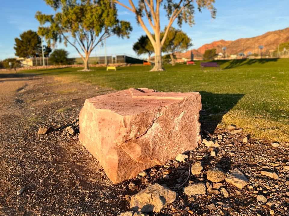 A large, rectangular Stone on a patch of dirt with scattered smaller rocks, surrounded by grass and trees in the background under a clear sky.