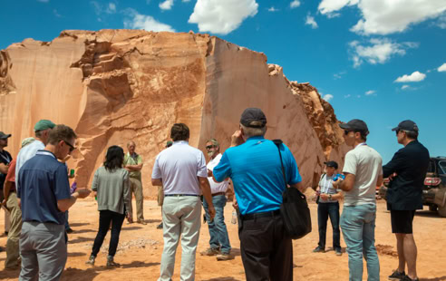A group of people standing on a sandy area in front of a large rock formation under a blue sky with clouds.