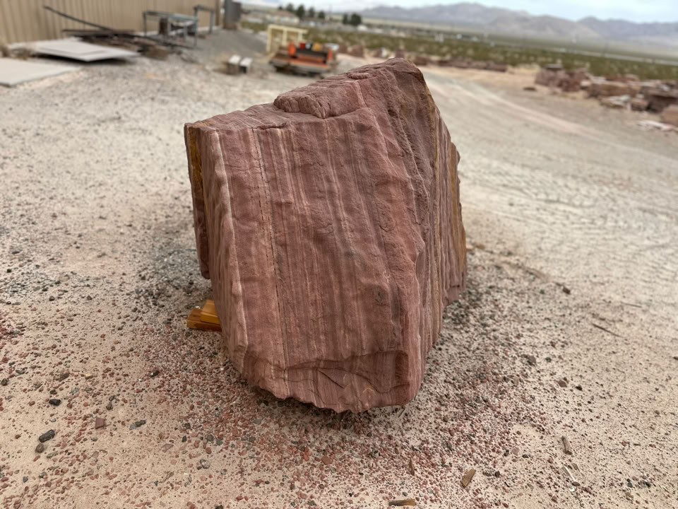 Large, rough, reddish-brown rock on a gravel surface with industrial equipment and distant mountains in the background.