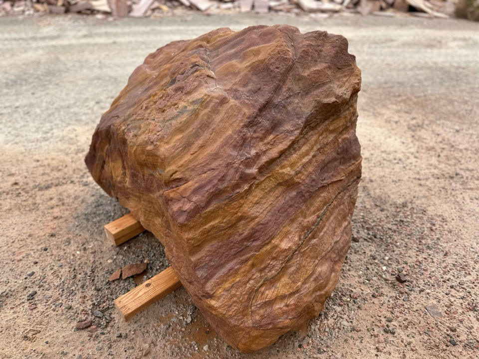 A large, textured brown Windswept Boulder resting on two wooden supports on a sandy surface.