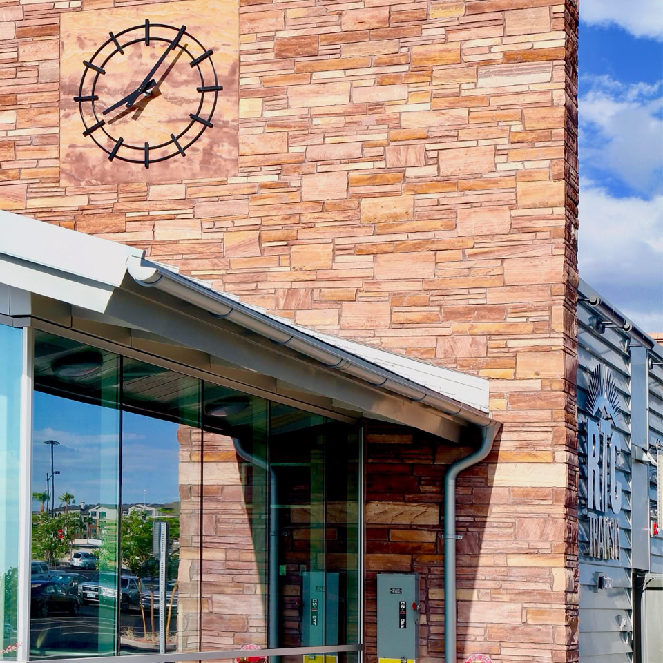 A building with a brick facade featuring a clock, large glass windows, and a metal roof, set against a blue sky with clouds.
