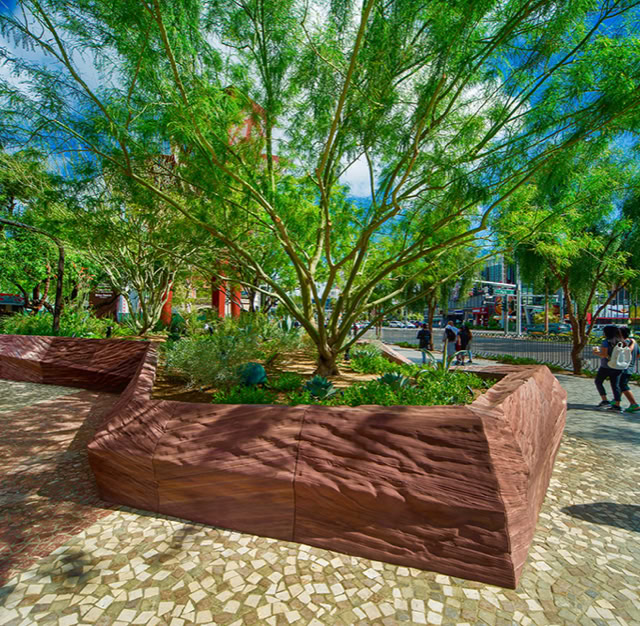 A landscaped area with a large tree and plants in a raised, angular stone planter, surrounded by a paved walkway and people walking in the background. @MGM THE PARK LAS VEGAS NV