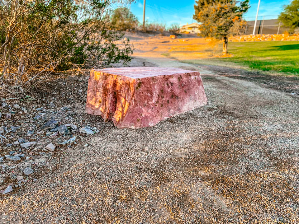 A large pinkish rock placed on a dirt path surrounded by grass and trees, with a clear blue sky in the background.