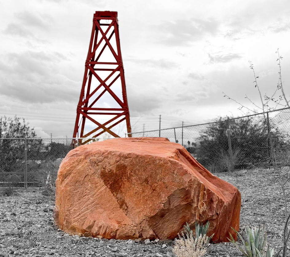 A large red rock in the foreground with a red metal tower structure in the background, set in a barren landscape with sparse vegetation and a cloudy sky.