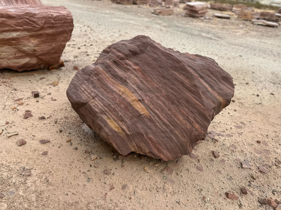 A large, textured, reddish-brown rock with visible layers sits on sandy ground.