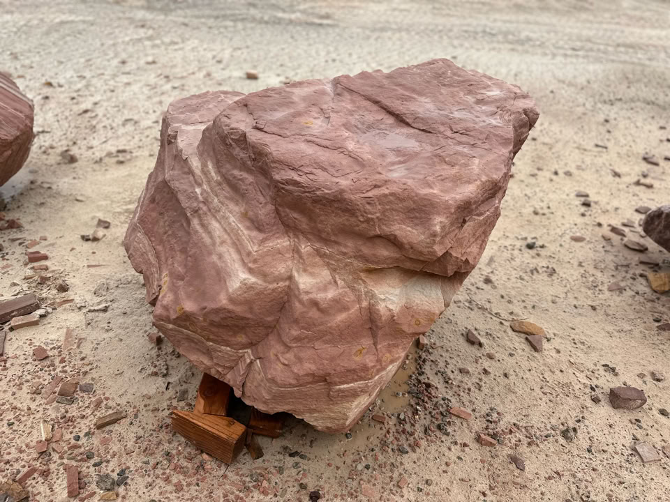 A large, rough, reddish-brown rock resting on sandy ground with small scattered stones around it.