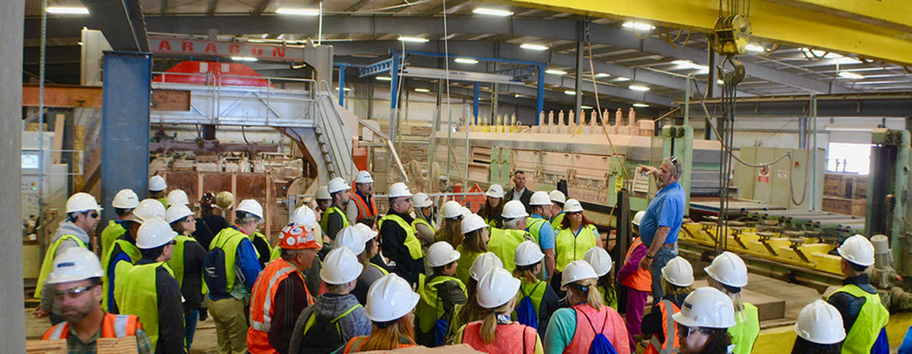 Teachers Workshop Tour people standing around in a factory with hardhats on as one man stands on the podium pointing out different equipment.