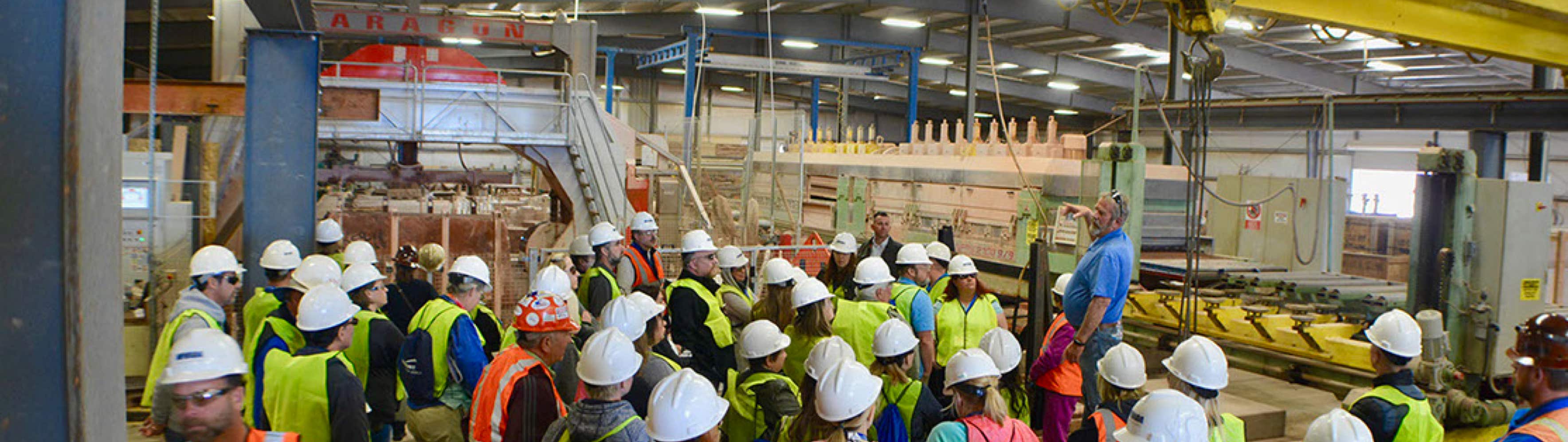 Teachers Workshop Tour people standing around in a factory with hardhats on as one man stands on the podium pointing out different equipment.