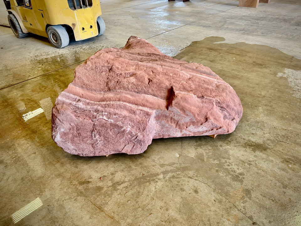 A large pink rock with white stripes on a concrete floor, with part of a yellow forklift visible in the background. And the boulder kinda looks like a T-Rex head without all the definition.