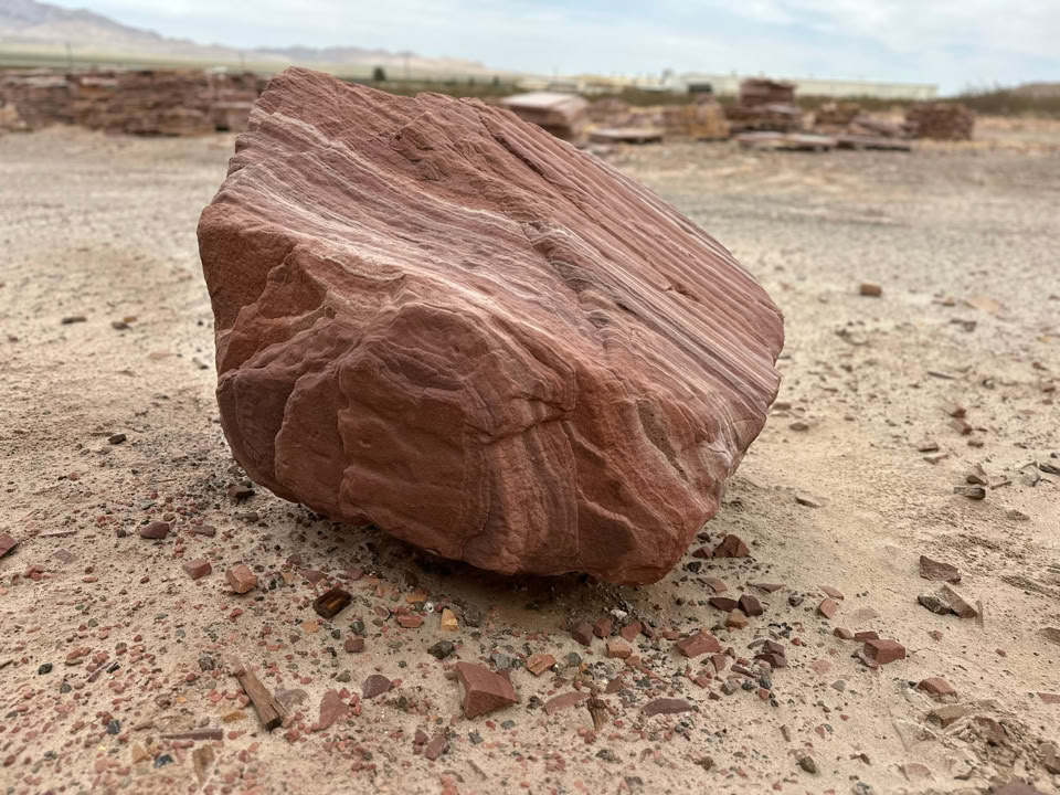 A large, reddish-brown rock with visible layers and striations sits on a sandy, rocky ground with a blurred background of more rocks and a cloudy sky.