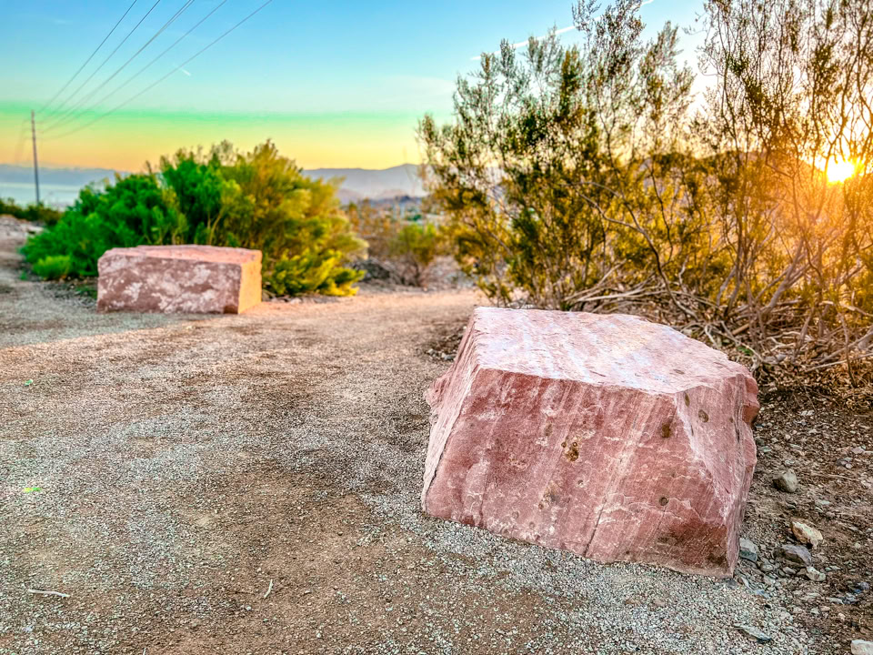 Two large pink stone blocks on a dirt path surrounded by bushes, with a sunset in the background.