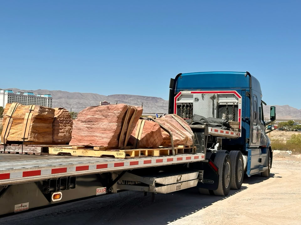 A blue semi-truck with a flatbed trailer carrying large stacks of stone slabs, set against a clear blue sky and distant mountains.
