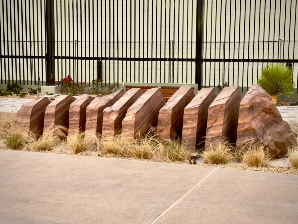 A row of large, sliced boulder arranged in a zigzag pattern, surrounded by dry grass, with a metal fence in the background. Located in Las Vegas, Nevada at the UNC trauma center by the emergency entrance