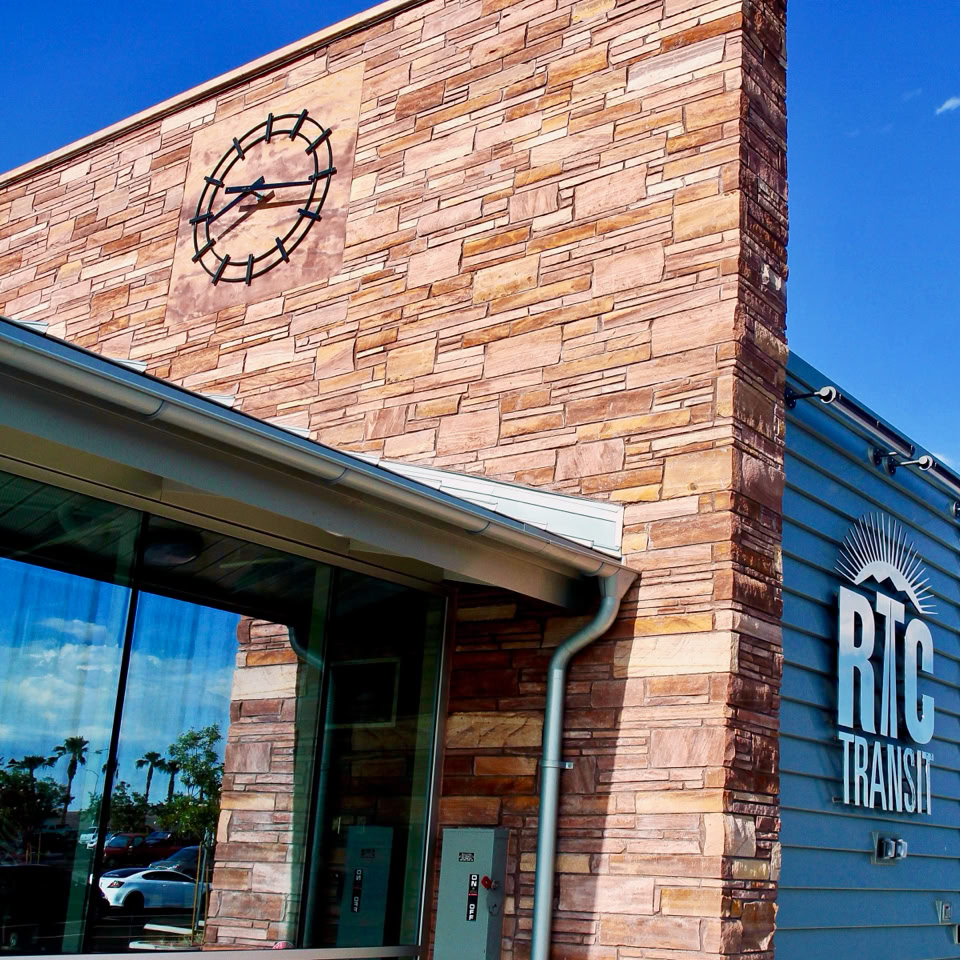 Building exterior with a brick wall and a clock, adjacent to a modern glass and metal structure with "RTC TRAILS" signage, under a clear blue sky.